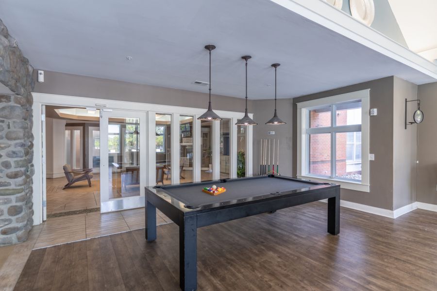 A modern game room with a black pool table, wooden floors, pendant lights, and large windows letting in natural light.