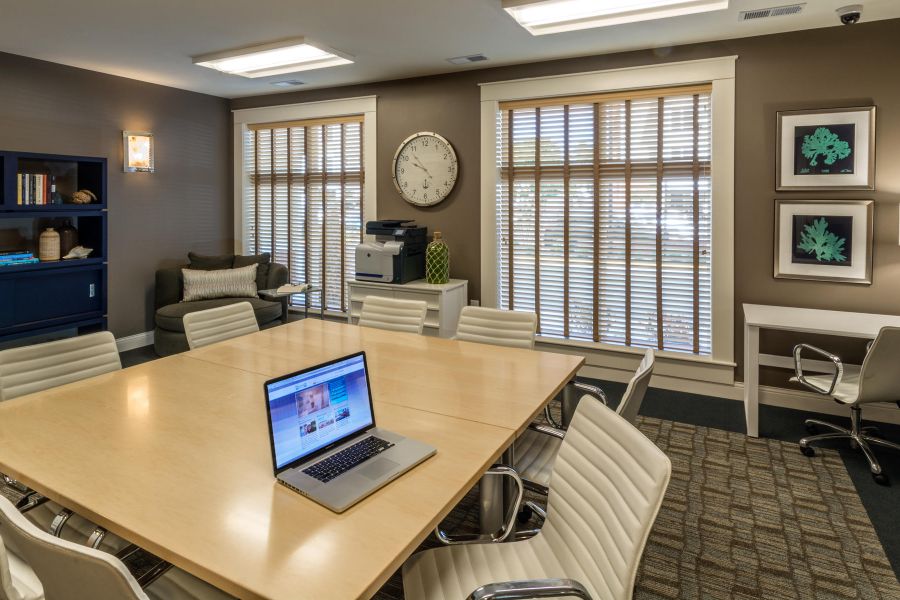 Modern office room with a large meeting table, laptop, chairs, bookshelf, desk, and windows with blinds.