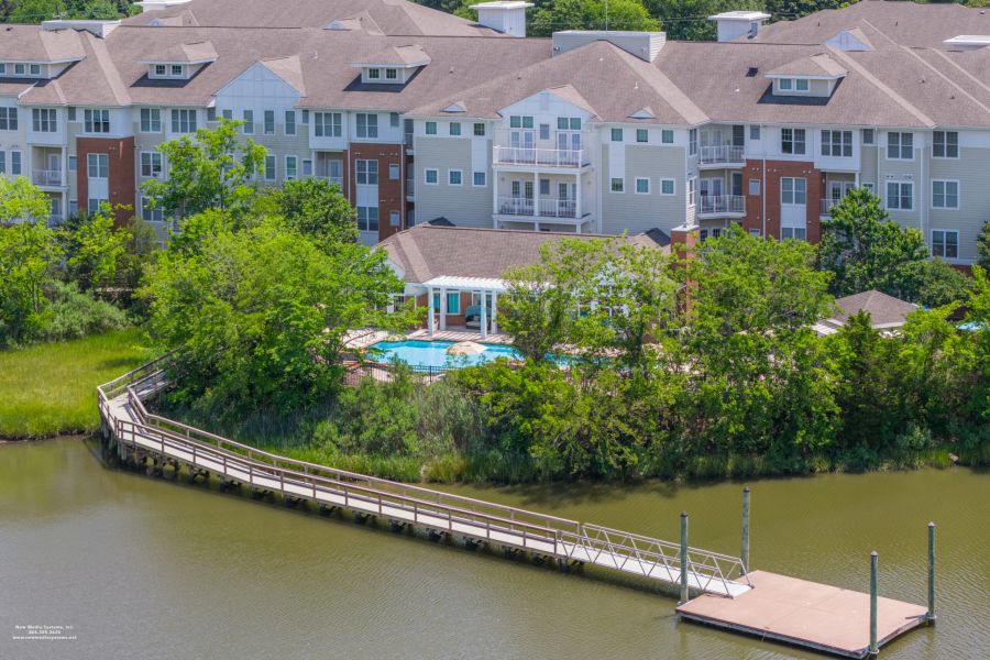 Apartment complex with a pool, surrounded by trees, and a wooden dock extending over a calm body of water.