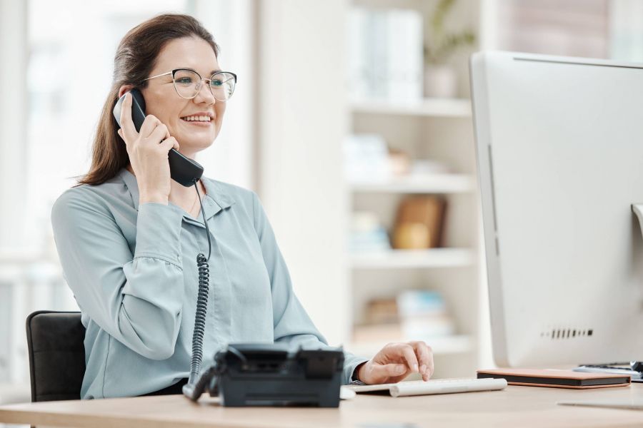 Attain on 5th apartment homes with Smiling woman at desk talks on the phone while using a computer in a bright office.