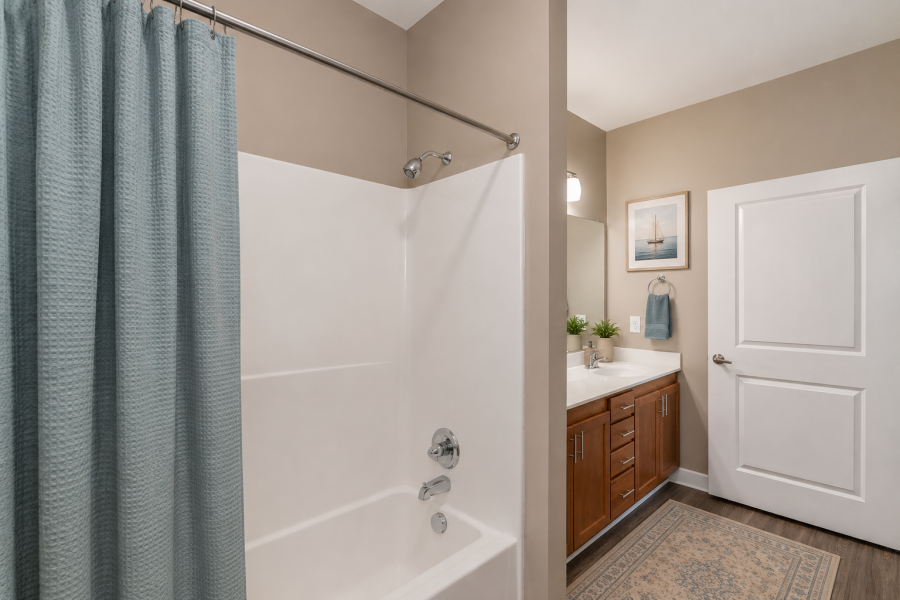Modern bathroom with a white tub, blue shower curtain, wooden vanity, and nautical wall art above a towel ring by the sink.