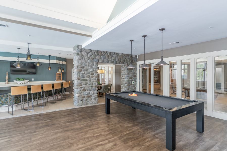 A modern game room with a black pool table, stone accent wall, bar seating, and large windows.