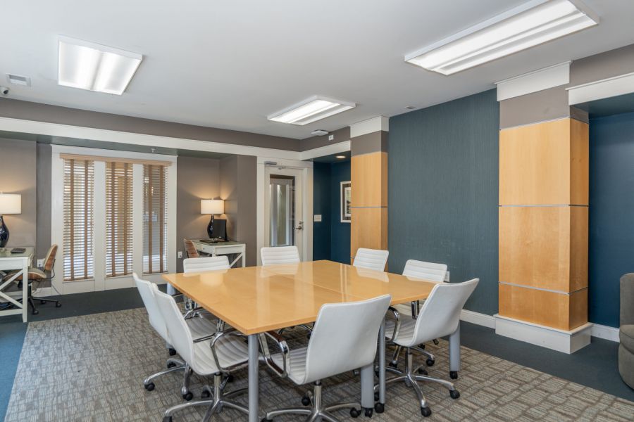 Modern conference room with a wooden table, white chairs, two desks, neutral walls, and large windows with blinds.