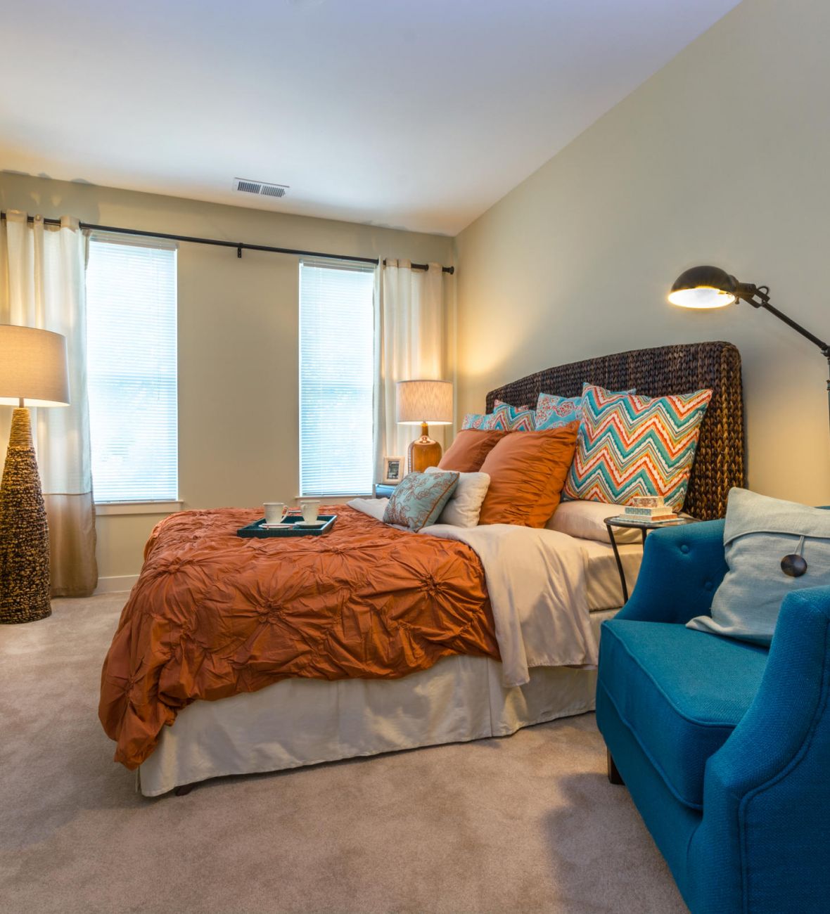 Cozy bedroom with orange bedding, blue armchair, wooden dresser, large mirror, and two tall windows.