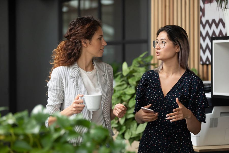 Two women stand indoors having a conversation; one holds a mug, the other holds a smartphone.