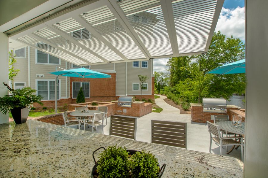 Outdoor patio with tables, chairs, grills, and blue umbrellas, viewed from a counter under a white pergola.