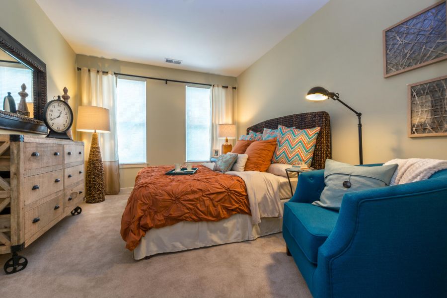 Cozy bedroom with orange bedding, blue armchair, wooden dresser, large mirror, and two tall windows.