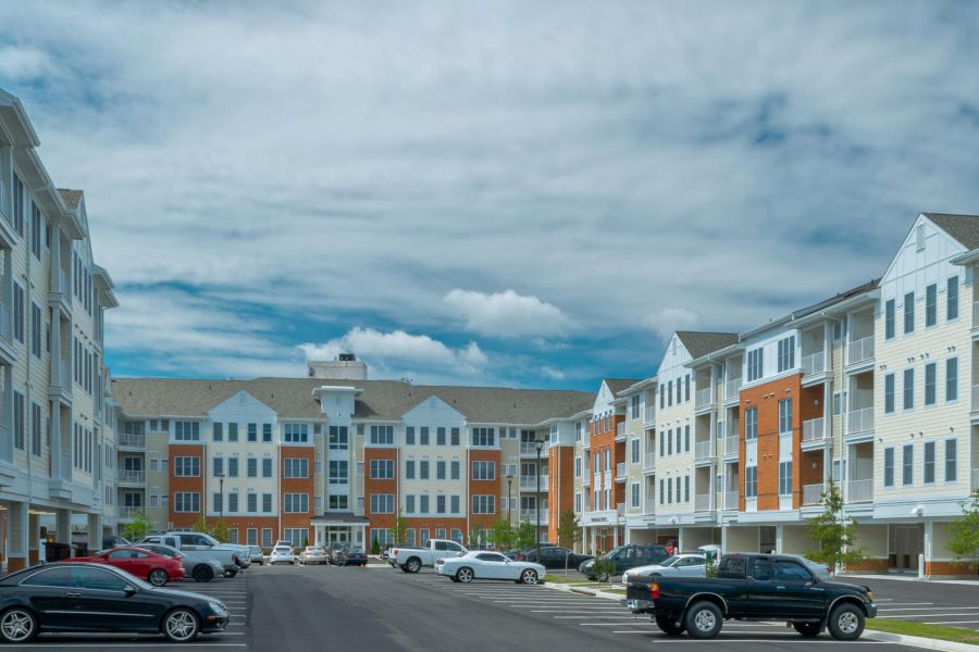 A wide view of an apartment complex with parked cars and a cloudy blue sky overhead.