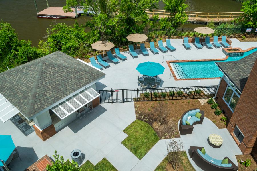 Aerial view of a pool area with lounge chairs, umbrellas, and a nearby river surrounded by trees.