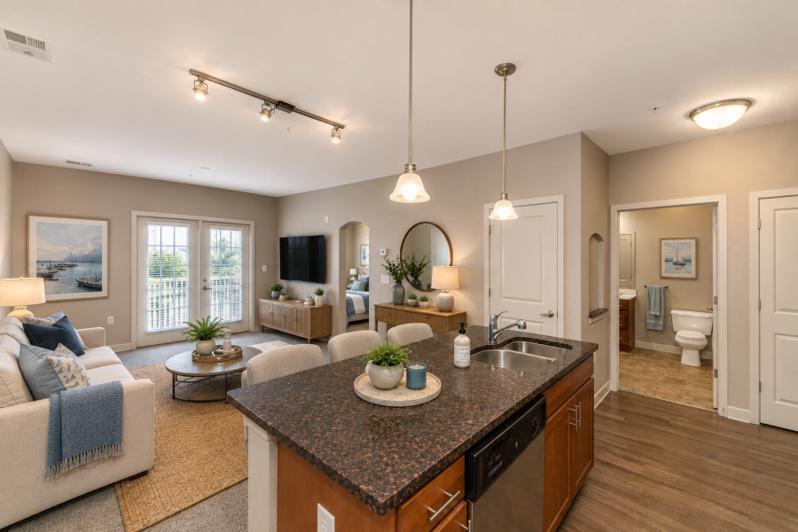 Open-plan living room with a kitchen island, sofa, TV, decor, and a view into a bathroom and hallway. Warm lighting and neutral tones.