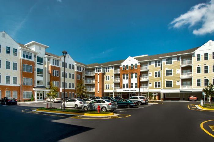 Four-story modern apartment complex with parked cars in front and clear blue sky above.